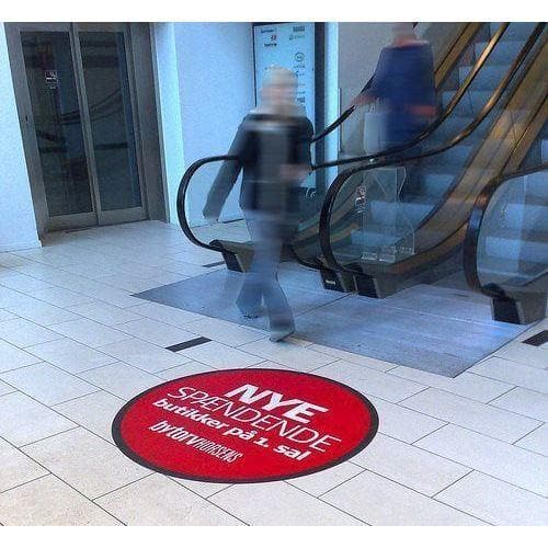 Rødt, rundt gulvklistermærke med hvid tekst på lyse fliser nær rulletrappe i offentlig rum.