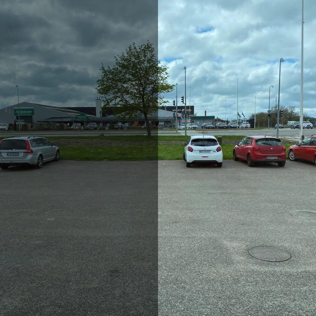 Parkeringsplads delt i to: venstre mørkere, højre lysere med biler, bygninger og træ i baggrunden.
