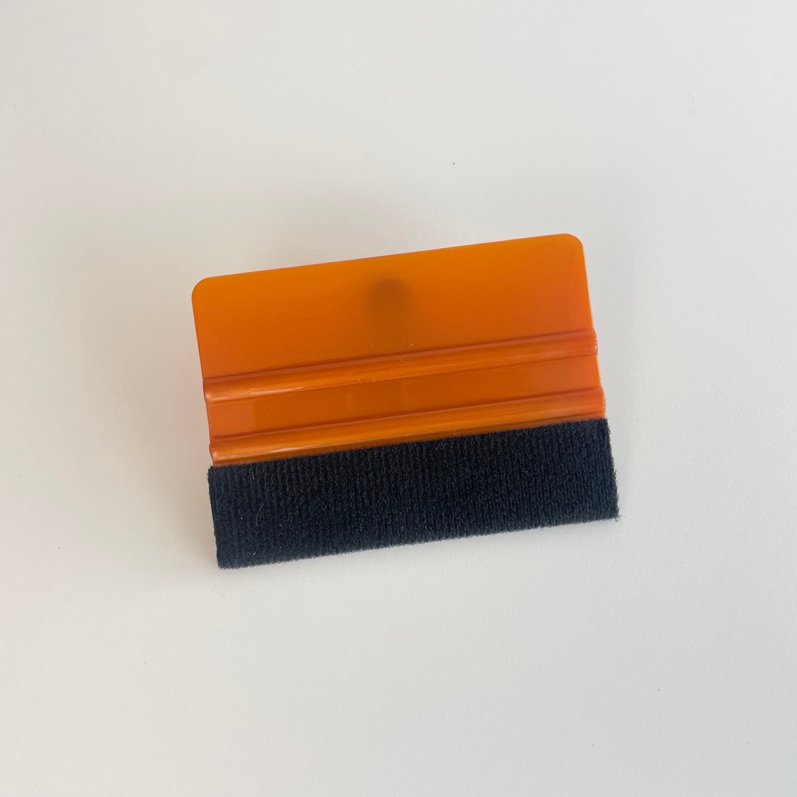 Lille orange skraber med sort filt. Rektangulær form, ca. 7x10 cm, let at håndtere.