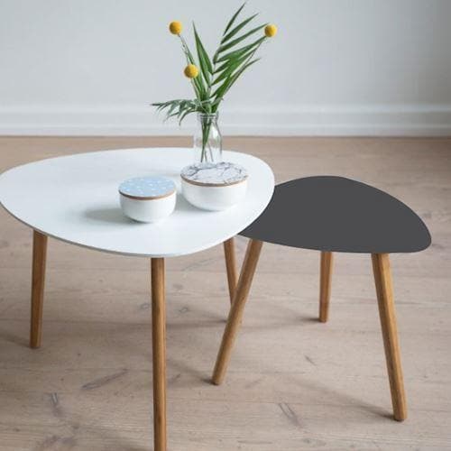 To små borde på trægulv; et mørkegråt, mat bord og et hvidt med vase og blomster.